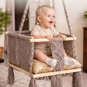 Baby sitting in white macrame baby swing with wooden base and cushion indoors